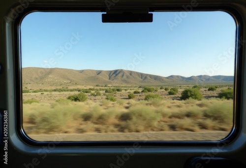View of arid landscape with rolling hills and sparse vegetation seen through a vehicle window, clear blue sky above and sunlight illuminating the scene