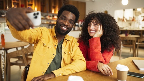 Man in yellow shirt taking selfie with smartphone while woman in red sweater laughs, sitting at cafe table. African American male and curly-haired female enjoying coffee and fun moment together indoor