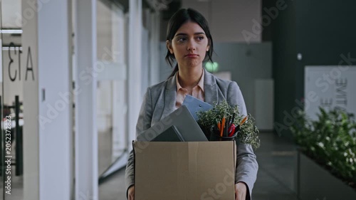 Worried Young Hispanic Female Employee Leaving Office Carrying Box Filled With Company Supplies And Plants After Job Termination. Empty Workspace Reflects A Business Concept.