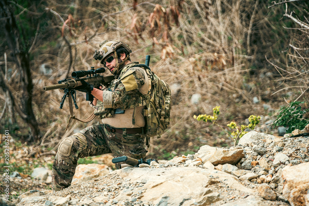 Fototapeta premium Armed soldier in camouflage kneels on a rocky hillside, aiming a scoped rifle during a field operation in dry, brushy terrain.