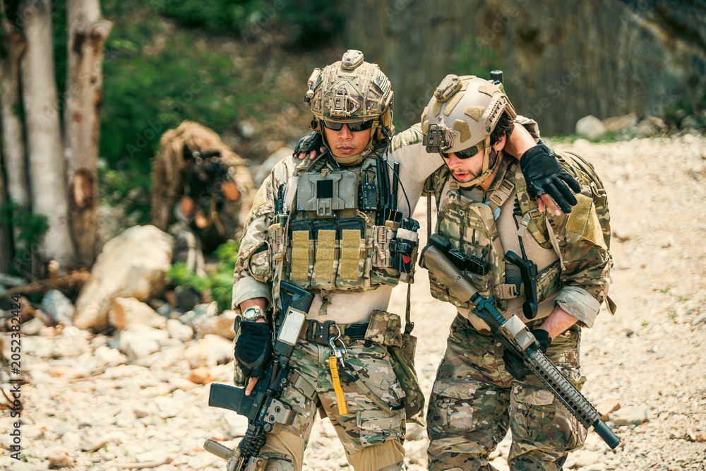Fototapeta premium Two soldiers in camouflage on rocky terrain; one supports an injured teammate as they move through the field, rifles and tactical gear visible during an outdoor operation.