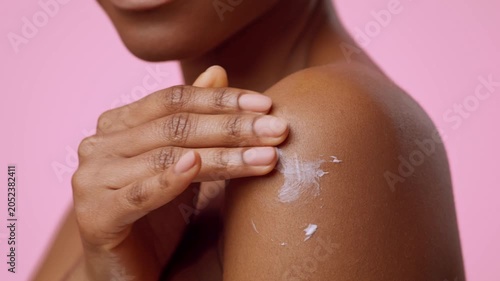 Focused Young Woman With Black Skin Applying Moisturizer On Her Shoulder In Front Of Pink Background. She Is Taking Care Of Her Skin With A Cream In A Home Setting