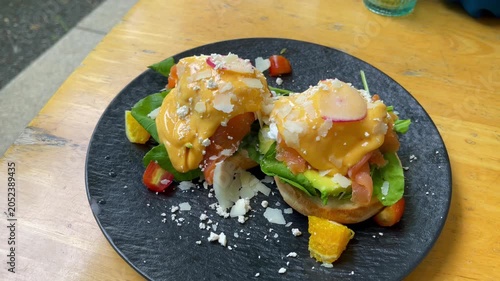 Gourmet eggs Benedict topped with smoked salmon, avocado, poached eggs, and creamy hollandaise sauce. Served on a black plate with fresh greens, parmesan shavings, and citrus garnish in a café setting