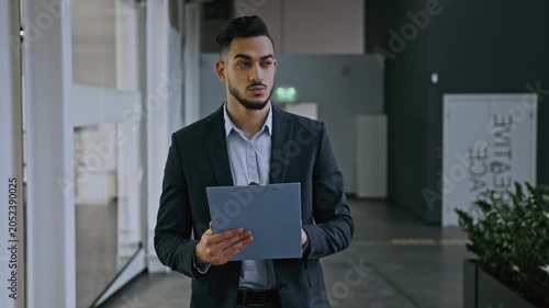 Focused Young Asian Man Dressed In Formal Attire Holding Clipboard While Standing In Office Setting. Professional Male Reviewing Documents In Modern Business Environment.