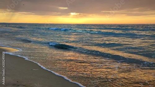 Sea waves with blue transparent water on sand of seashore on sunset and sunrise. Yellow reflection of sunlight on surface of sea waves. Sky with dark rain clouds, horizon line and sea waves on sand.