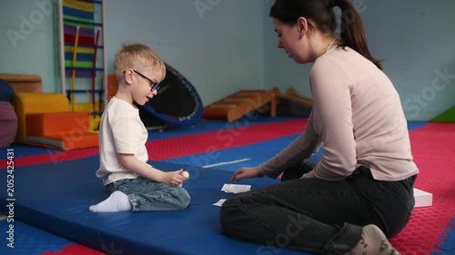 A small child wearing glasses sniffs and guesses smells while playing an educational game. on a blue mat in a bright indoor play space with various soft play structures.