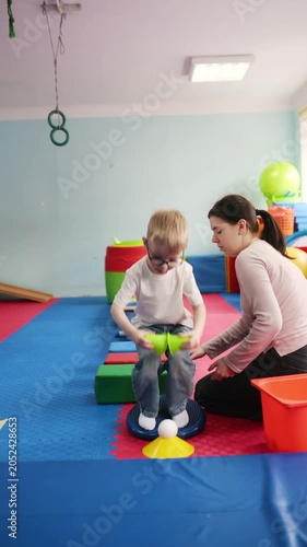 Young boy wearing glasses walks on a soft play structure while holding cups, with a woman assisting him in a colorful indoor play area