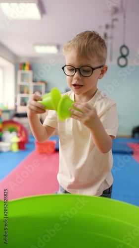 Young boy wearing glasses carries a green cup while walking in a vibrant playroom with soft mats, toys, and shelves filled with colorful items