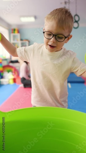 Young boy wearing glasses carries a green cup while walking in a vibrant playroom with soft mats, toys, and shelves filled with colorful items