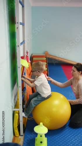 Young boy wearing glasses balances on a yellow exercise ball as a female caregiver supports him in a vibrant indoor playroom filled with equipment