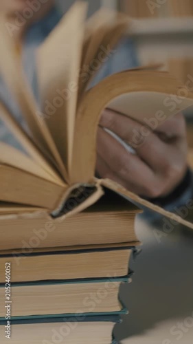 A man is reading a book, examining the pages carefully. Several books are stacked beneath his hands, indicating a moment of focused study or research.