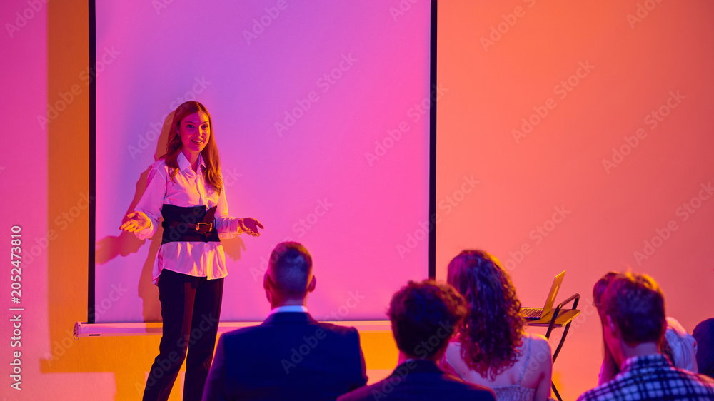 © Lustre Art Group - Female presenter speaking to audience beside blank screen in neon seminar room. Concept of business seminar, workshop training, public speaking, leadership and corporate education. © Lustre Art Group - Female presenter speaking to audience beside blank screen in neon seminar room. Concept of business seminar, workshop training, public speaking, leadership and corporate education.