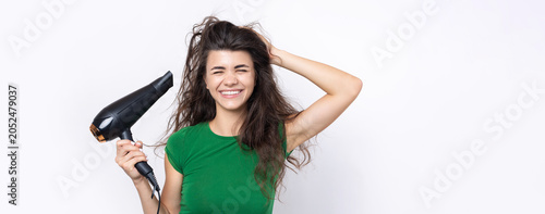 A cute young girl dressed in a green top dries her beautiful long silky hair with a hair dryer against a white background.