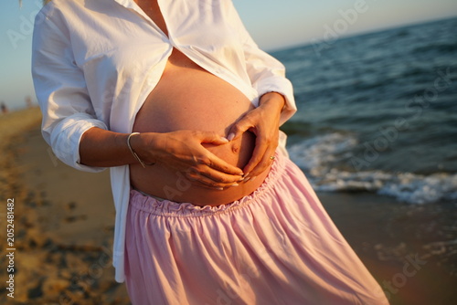 Foto Pregnant woman stands on the beach, forming a heart shape on her bare belly with her hands, symbolizing love and anticipation
