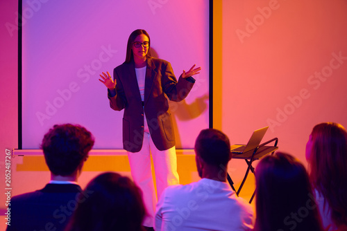 Konstfotografi Serious female speaker addressing audience with open hands in neon seminar setting