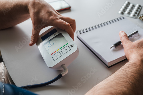 Obraz na płótnie Close-up of a mature man's hands on a digital home blood pressure monitor while writing the reading in a notebook illustrating hypertension self-monitoring and health tracking at home