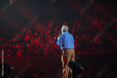 Older male speaker on stage with microphone, large seated audience in red lighting, live event atmosphere, public speaking and performance.