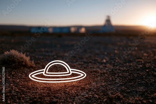 A remote desert landscape at sunset featuring a glowing outline of a UFO on the ground with distant structures