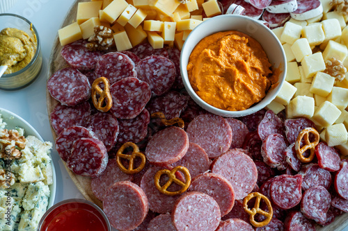 Φωτογραφία A round sharing board with salami slices cheese cubes pretzels walnuts and a red pepper dip on a white table a festive charcuterie platter for entertaining