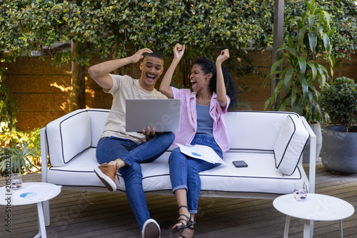 Diverse couple cheering and laughing while reviewing charts on patio sofa with laptop, plants