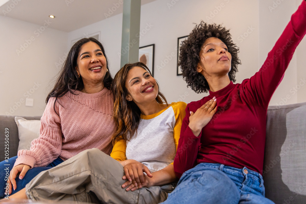 Naklejka premium Diverse female friends sitting on gray couch in living room holding hands taking selfie with phone