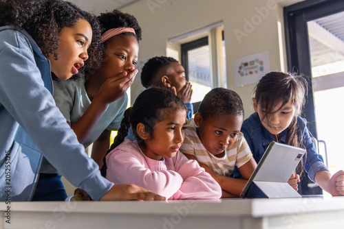 Foto Diverse group of school-age children leaning around tablet in classroom, holding