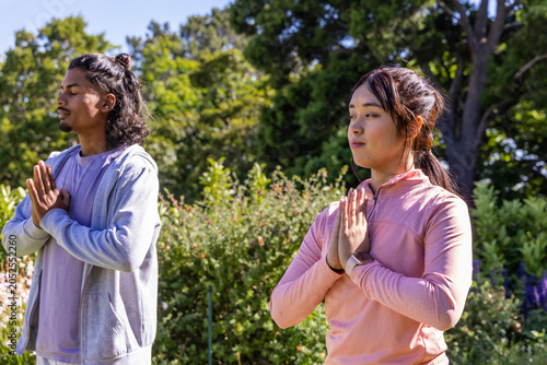Diverse couple meditating with palms at chest in park wearing athletic gear and smartwatch