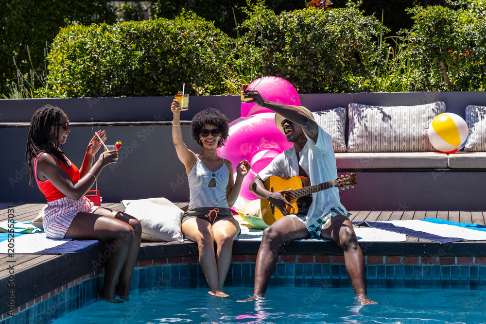 Naklejka premium African American friends sitting in swimwear at pool, playing guitar and holding drinks by flamingo