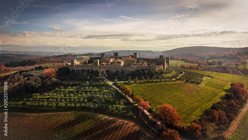 An aerial view of a medieval walled village perched on a Tuscan hill, surrounded by golden vineyards and olive groves during a serene late afternoon sunset in Italy.