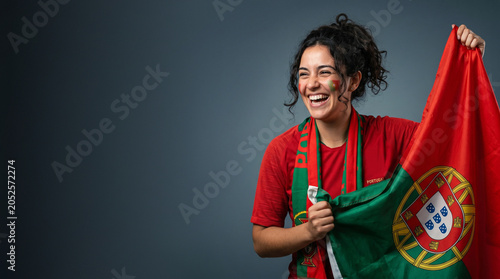 Cheerful Portugal football fan holding national flag, happy female supporter in red jersey with face paint laughing, cheering for Portuguese soccer victory isolated on studio background