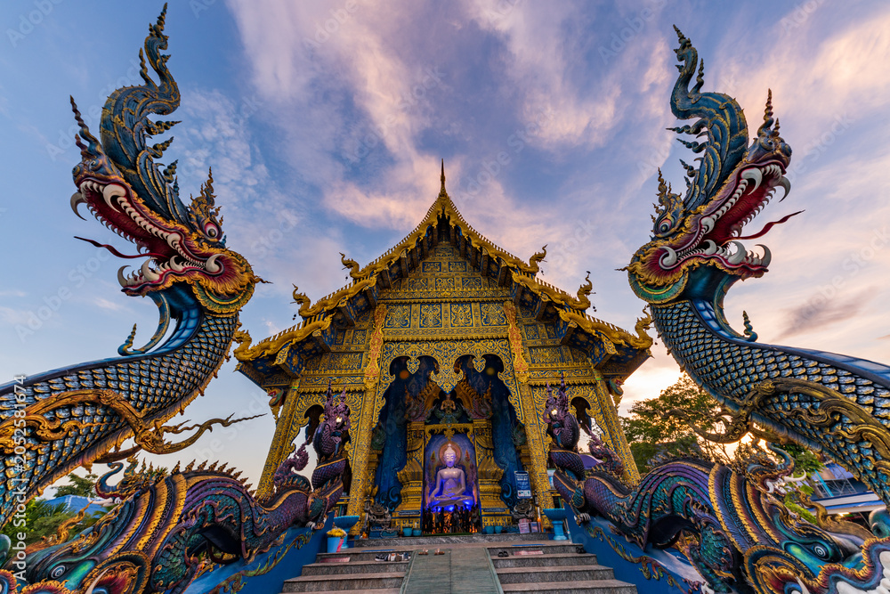 Fototapeta premium Majestic symmetrical view of giant blue Naga serpent statues guarding the entrance of the beautiful Blue Temple or Wat Rong Suea Ten at sunset, Chiang Rai, Thailand.