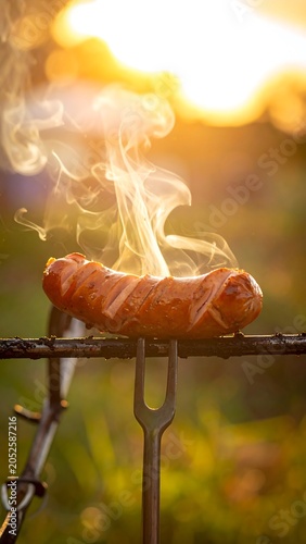 A grilled sausage sizzles on a metal grill with smoke rising at sunset