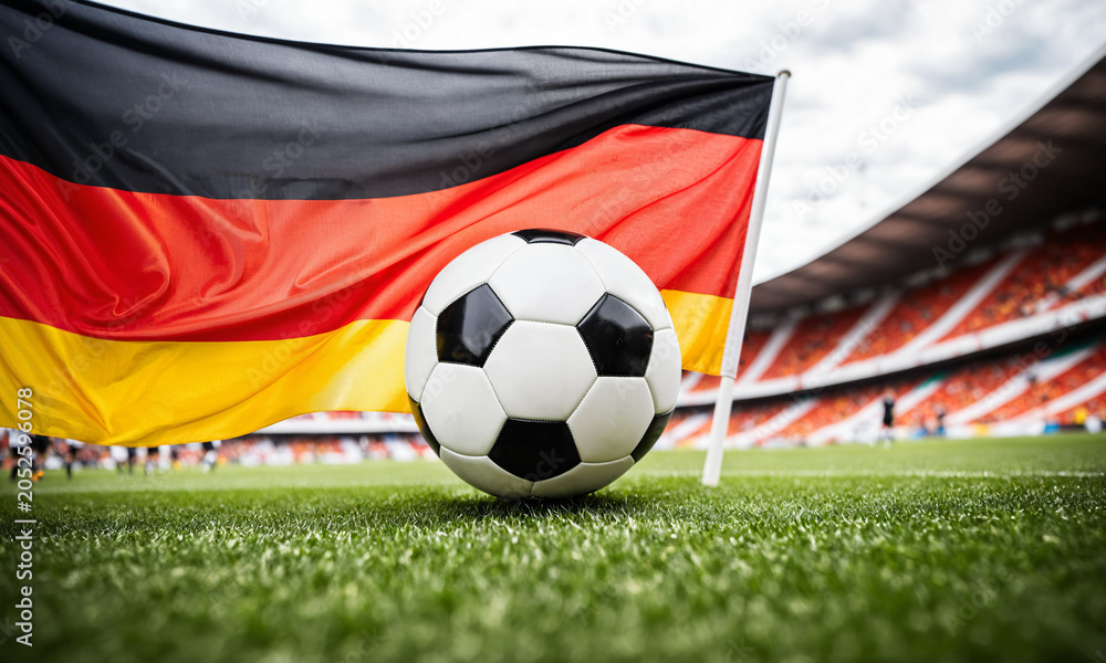 Naklejka premium soccer ball on green grass field in front of a large waving german national flag inside a stadium