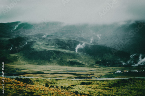 Misty geothermal valley in Iceland with volcanic mountains, green hills, rising steam, and dramatic cloudy atmosphere.
