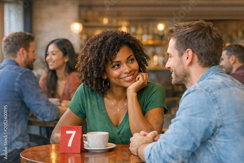 Couple smiling and talking in cozy cafe. Woman with curly hair wearing green top. Man in denim shirt leaning forward. Table with coffee cup and red table number 7. Background shows other patrons