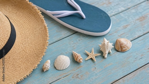 Seashells arranged with a straw hat and flip-flop on a blue plank, evoking a minimalist beach vibe.