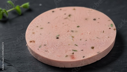 Close-up of pale pink liverwurst slice with herbs on slate.