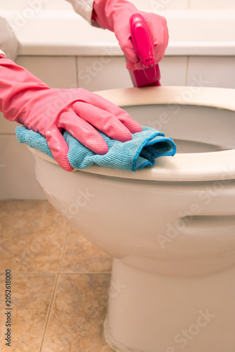 Pink gloves cover hands. Blue cloth wipes toilet seat. Person sprays cleaner on toilet. Focus on cleaning hygiene. Bathroom tiles in background. Cleanliness concept for home maintenance