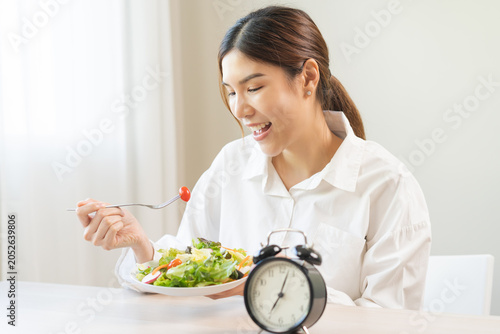 Intermittent fasting concept, close up clock, asain young woman, girl diet eating ketogenic low carb, fresh green vegetable salad on plate. Eat food healthy first meal in morning on table at home.