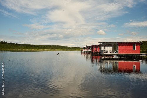 A solitary red wooden cabin built on stilts extends over a glassy lake, its bright facade and railing perfectly mirrored in the still water. The scene shows a