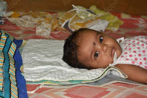 Indian little baby girl portrait in a white bodysuit on the bed at home, five month old newborn baby laying down on bed and rejoices.