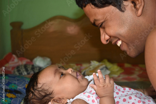 Parents playing with baby girl while lying on wooden bed in cozy home setting. side view showing joyful bonding, parenting moments love for newborn in Indian modern family lifestyle.
