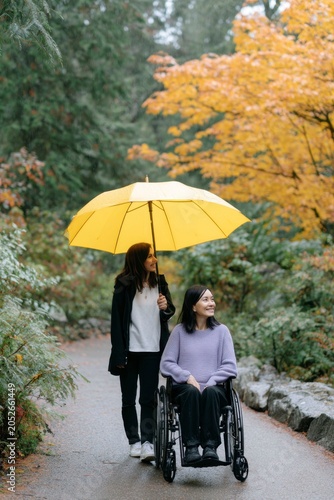 an adult woman in casual , holding up a yellow umbrella over her friend who is sitting in a wheelchair while walking along a path through a park during rain, with trees covered in autumn leaves.