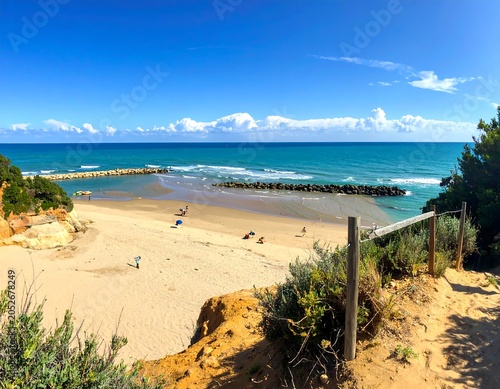 Sunny coastal landscape featuring a sandy beach, calm ocean, and a small jetty. Clear blue skies. Small beachgoers are visible