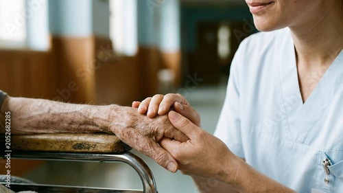 A healthcare worker in blue scrubs tenderly holds a senior's wrinkled hand on a wheelchair. This close-up conveys deep reassurance, gentle support, and dedicated medical care.