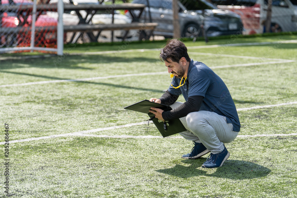 Naklejka premium Professional soccer coach squatting on green turf field writing strategy on clipboard outdoor, football trainer or referee preparing tactical notes during match training session