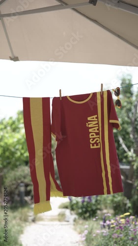 Soccer fan shirt, scarf and sunglasses in red and yellow national colors of Spain hanging on line under parasol outdoors in garden on sunny day for drying, background blurred
