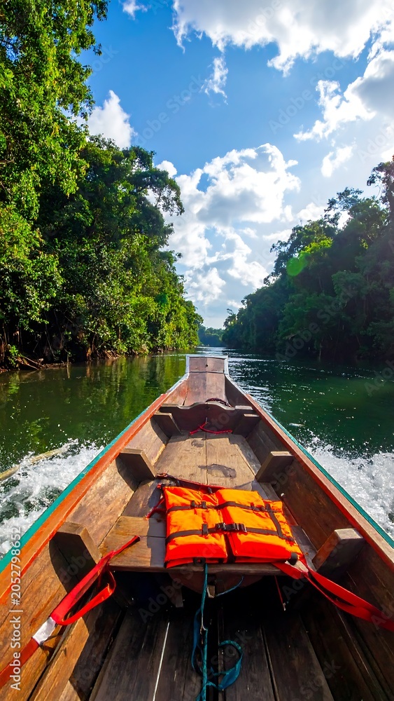 Fototapeta premium Wooden boat navigating river surrounded by lush green trees and blue sky