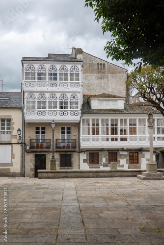 Traditional white glass balconies and an ancient stone cross in the historic center of Viveiro, Galicia, Spain under a cloudy sky.