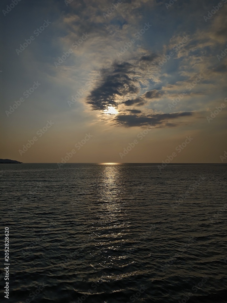 Obraz premium Dramatic sunset over the dark ocean with the sun hiding behind clouds and reflecting on the water surface.